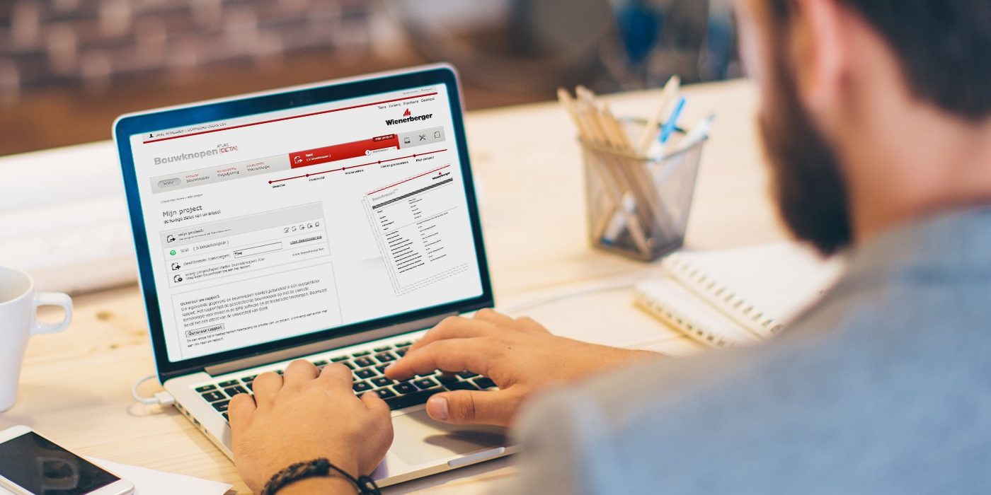 Young man working at modern office space, working on project. Sitting at the table and using laptop. Brick wall in background. Over the shoulder view.