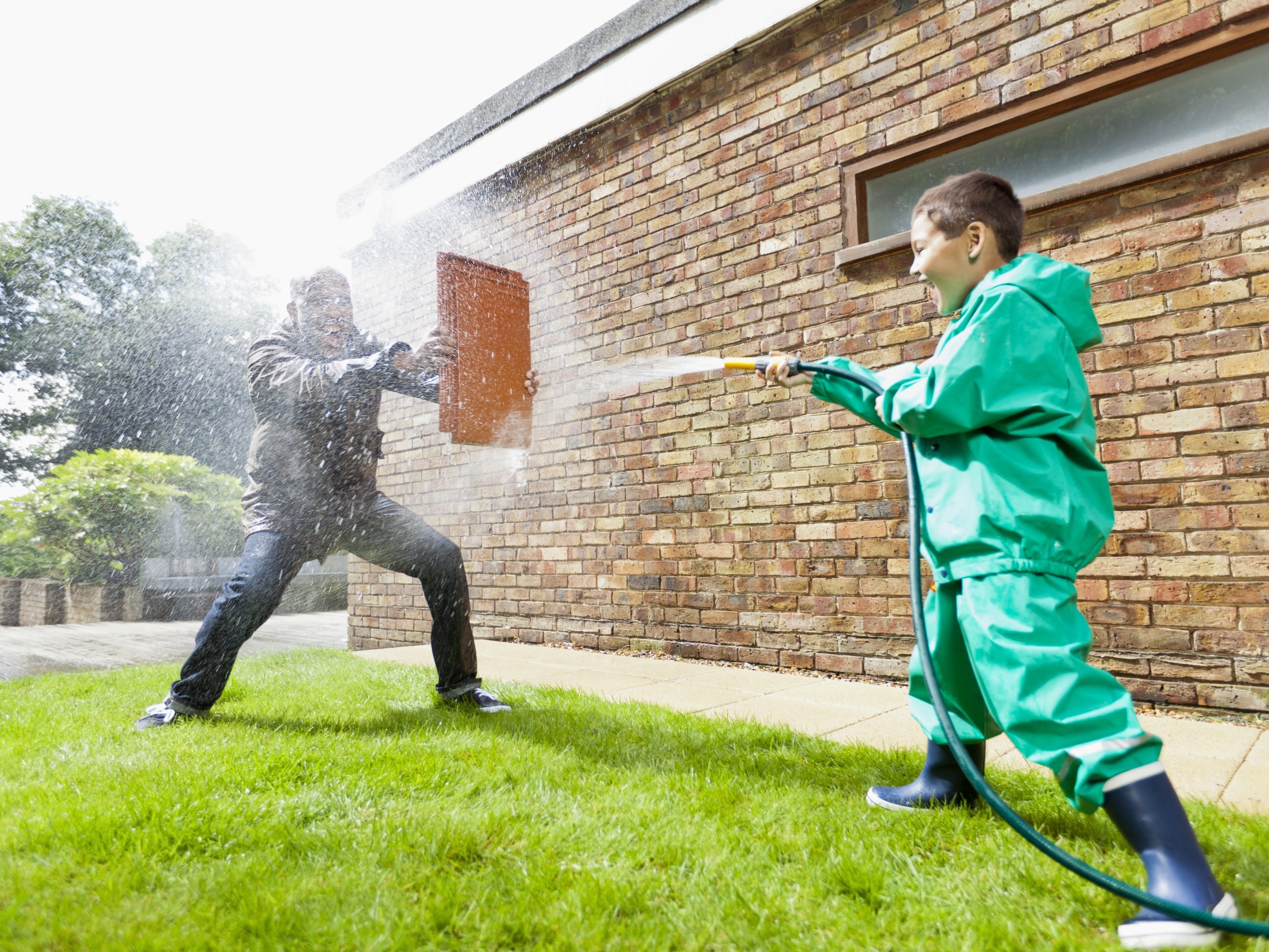 Boy hosing man holding a clay roof tile for protection in front of brick-built façade