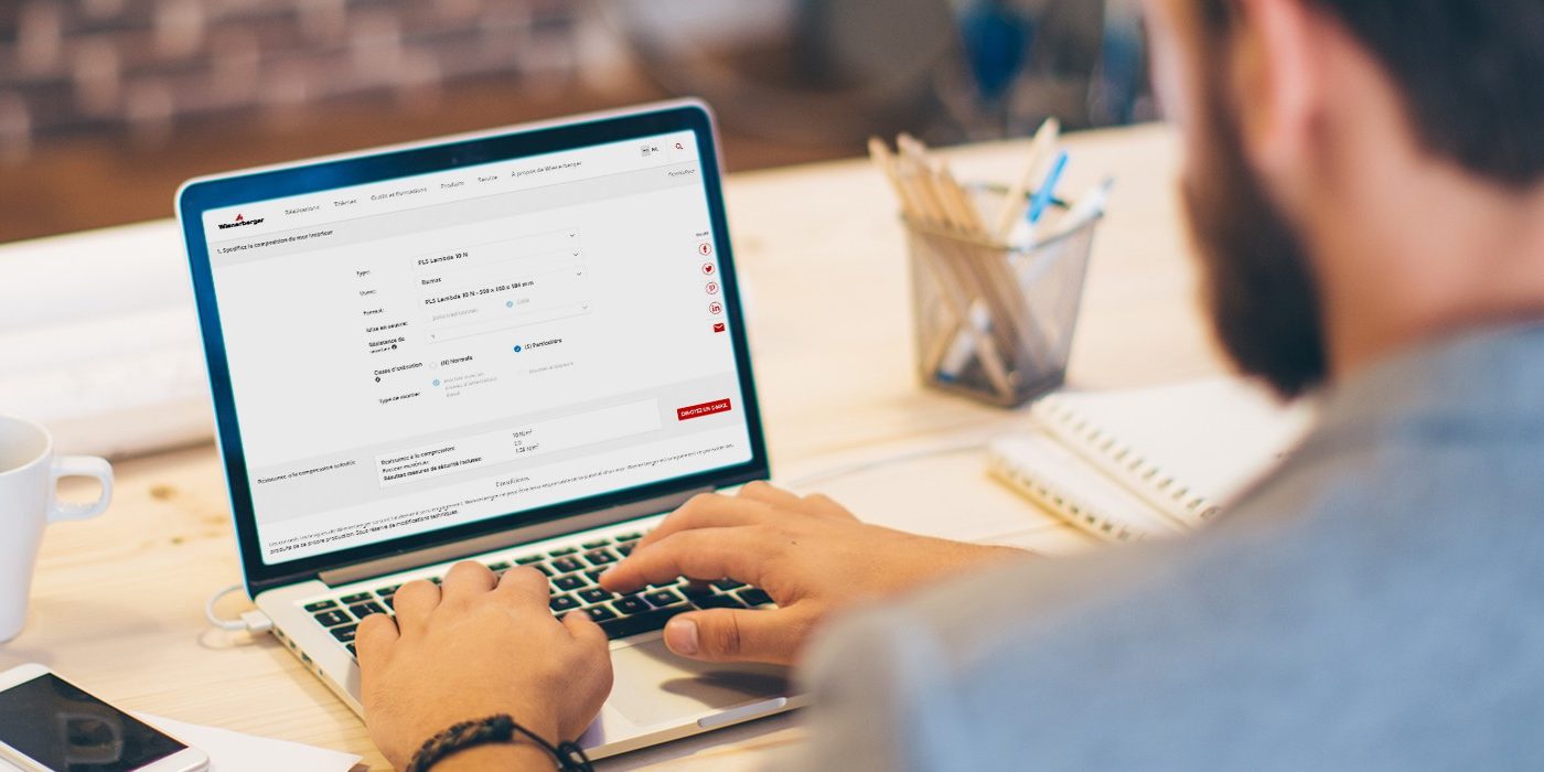 Young man working at modern office space, working on project. Sitting at the table and using laptop. Brick wall in background. Over the shoulder view.