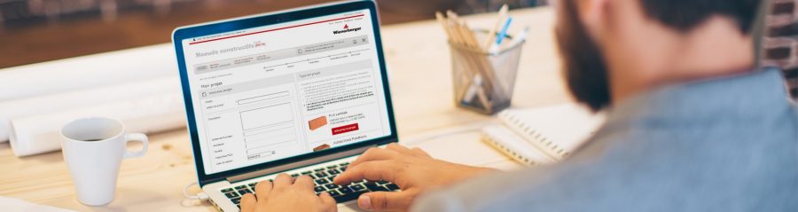 Young man working at modern office space, working on project. Sitting at the table and using laptop. Brick wall in background. Over the shoulder view.