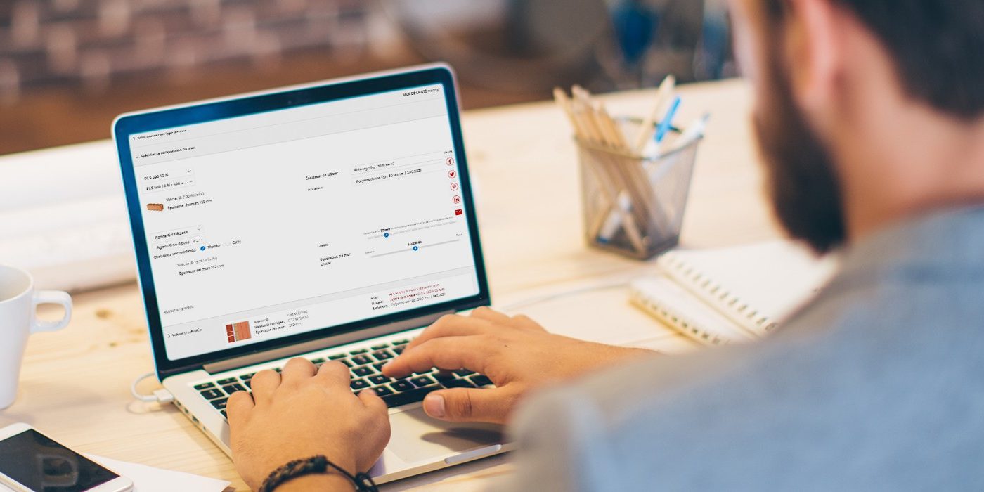 Young man working at modern office space, working on project. Sitting at the table and using laptop. Brick wall in background. Over the shoulder view.