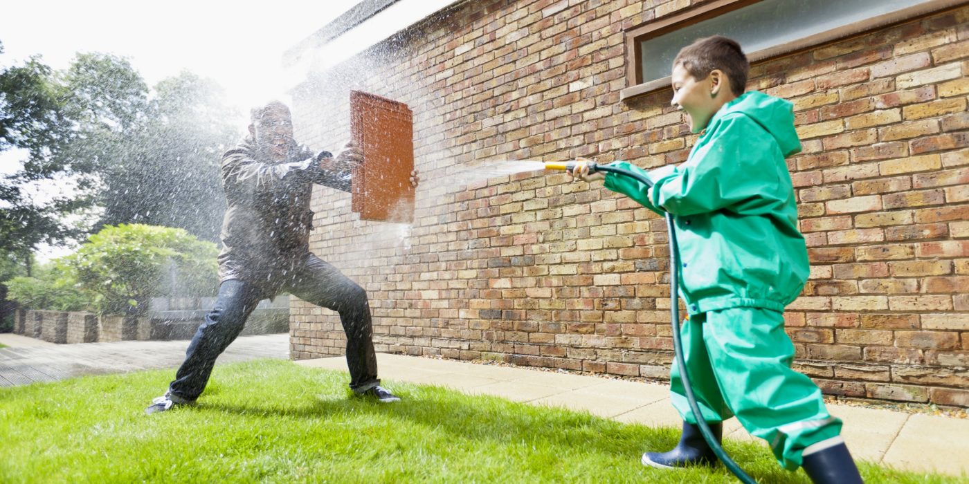 Boy hosing man holding a clay roof tile for protection in front of brick-built façade