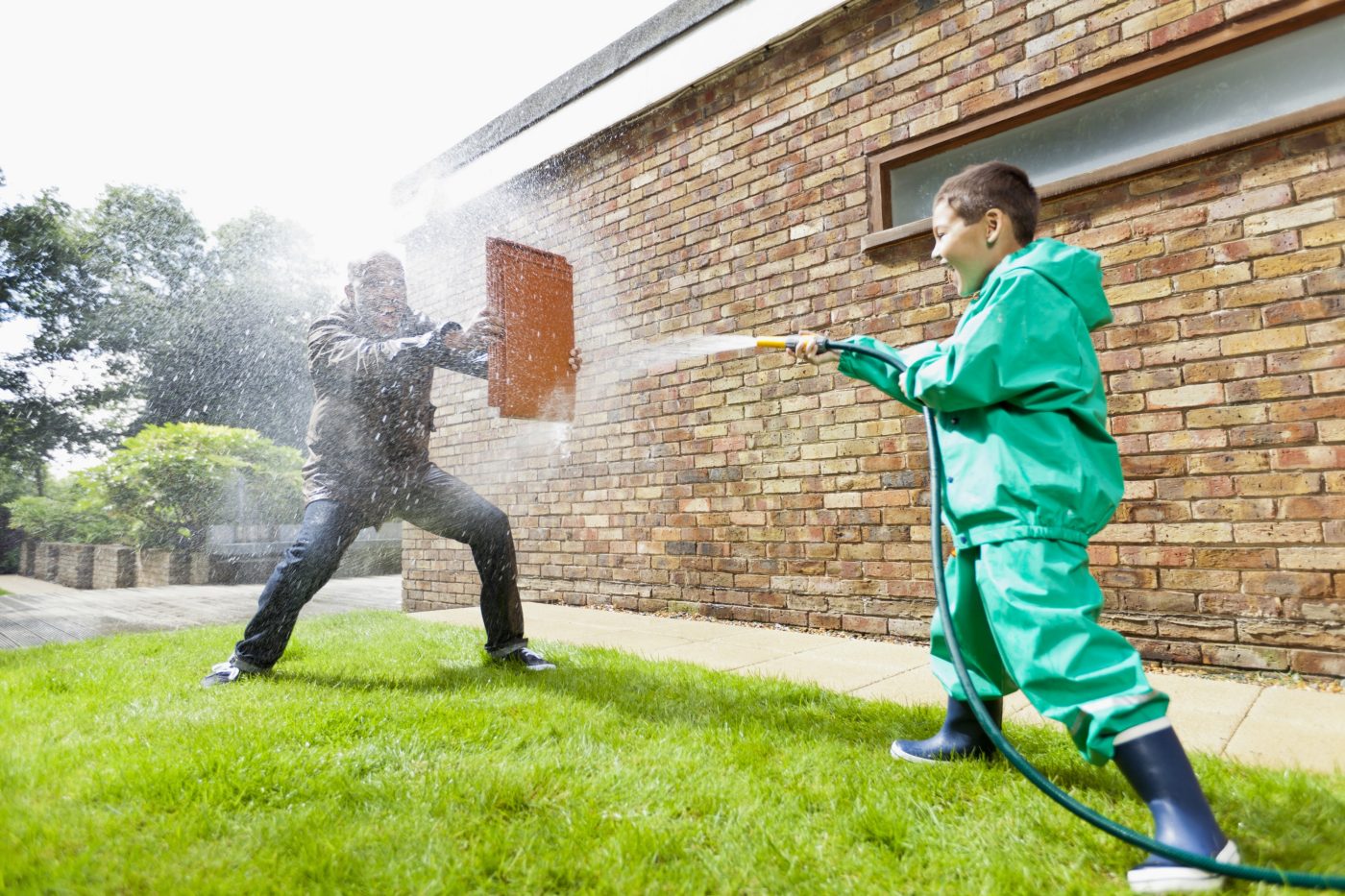 Boy hosing man holding a clay roof tile for protection in front of brick-built façade