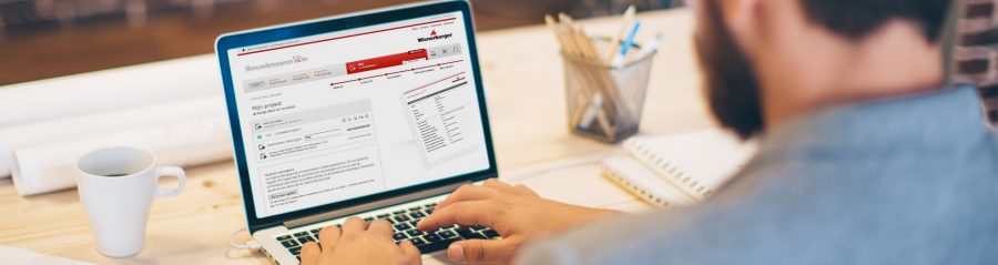 Young man working at modern office space, working on project. Sitting at the table and using laptop. Brick wall in background. Over the shoulder view.